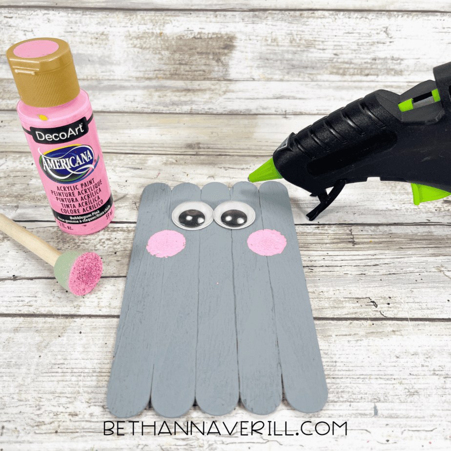 Grey painted popsicle sticks with googly eyes glued on and pink cheeks painted.