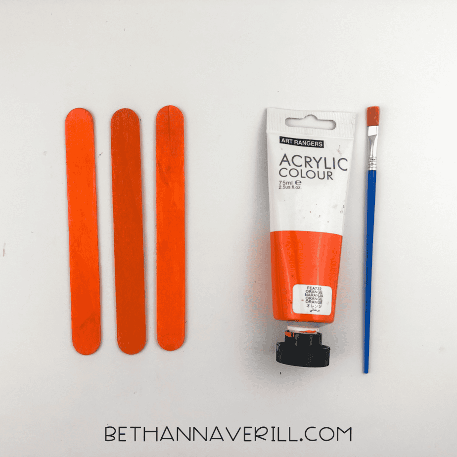 Three craft sticks painted orange next to a tube of acrylic paint and brush.