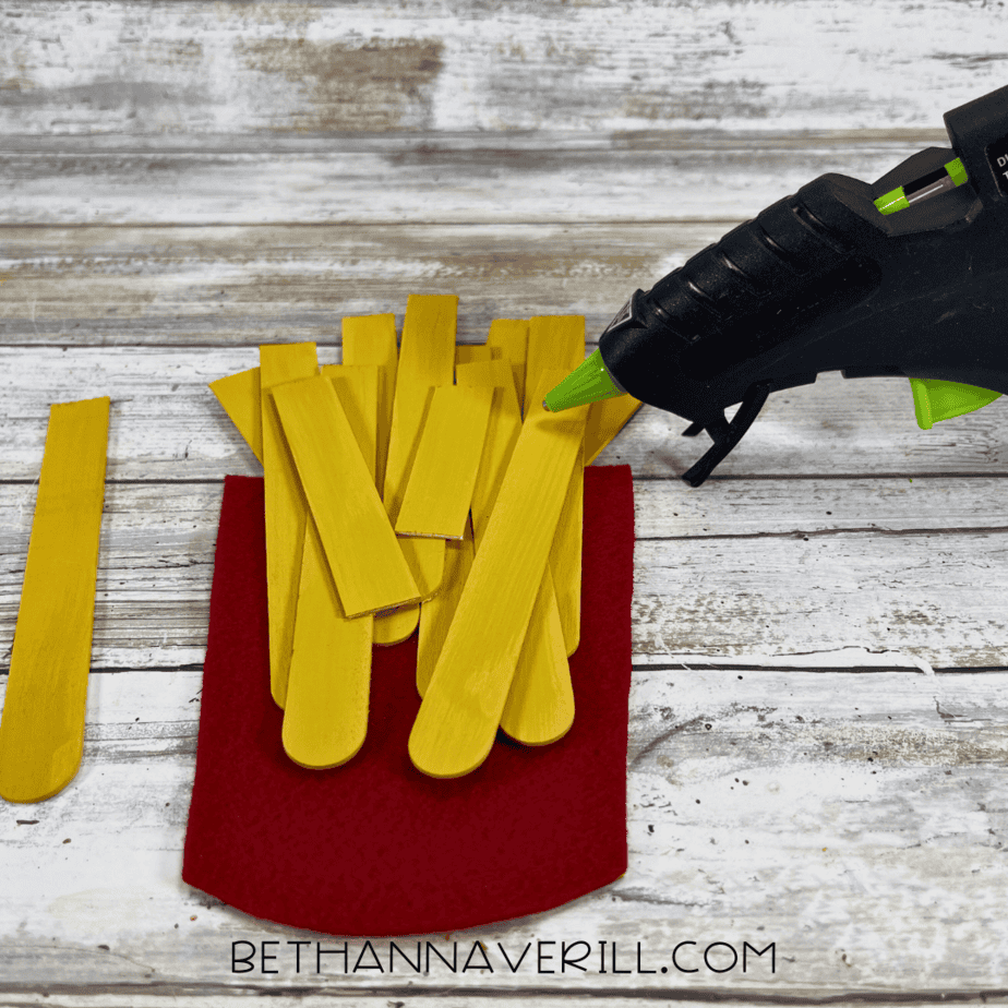Yellow painted craft sticks being glued inside red felt container with hot glue gun.