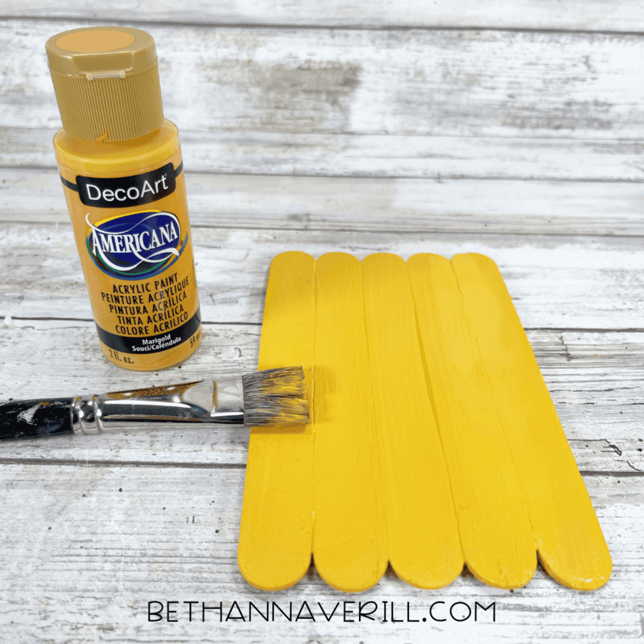Popsicle stick base painted yellow with paintbrush and yellow paint bottle.