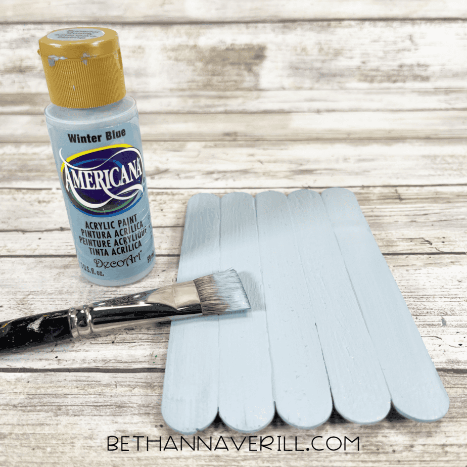 Popsicle sticks painted light blue with a paintbrush and bottle of acrylic paint.