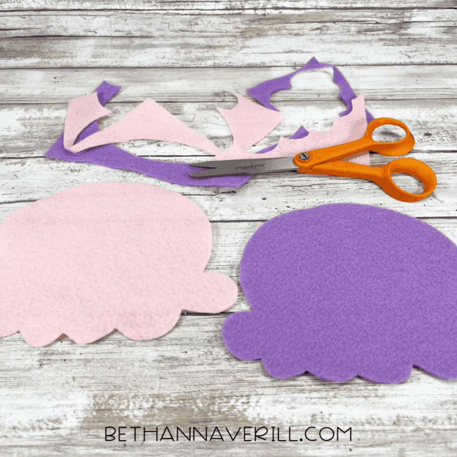 Two ice cream scoop shapes cut out of pink and purple felt, with scraps and scissors nearby on a wooden surface.