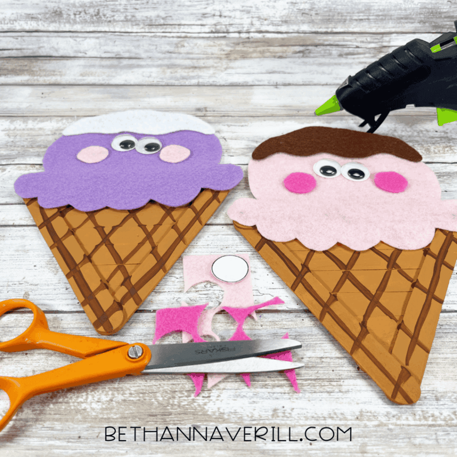 Two felt ice cream faces on cone-shaped craft stick bases, with scissors, glue gun, and cut pink cheeks.