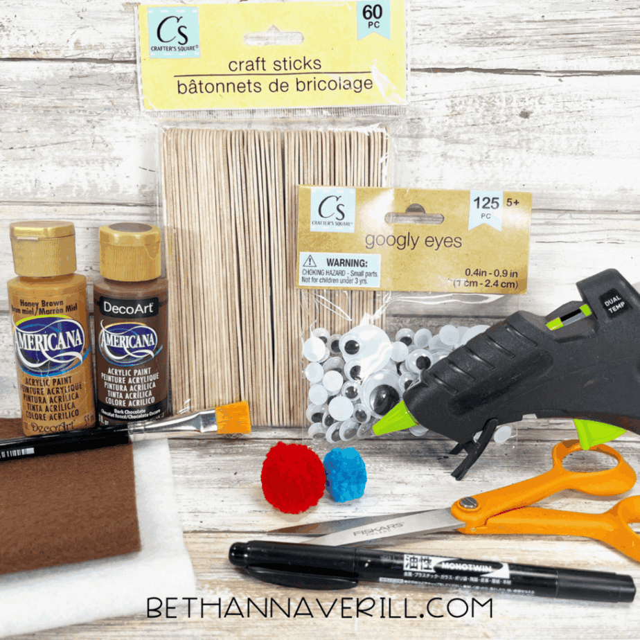 A collection of crafting materials including craft sticks, googly eyes, acrylic paint in brown and tan, two pom-poms, brown and white felt sheets, scissors, a hot glue gun, and a black marker, all arranged on a rustic wood background.