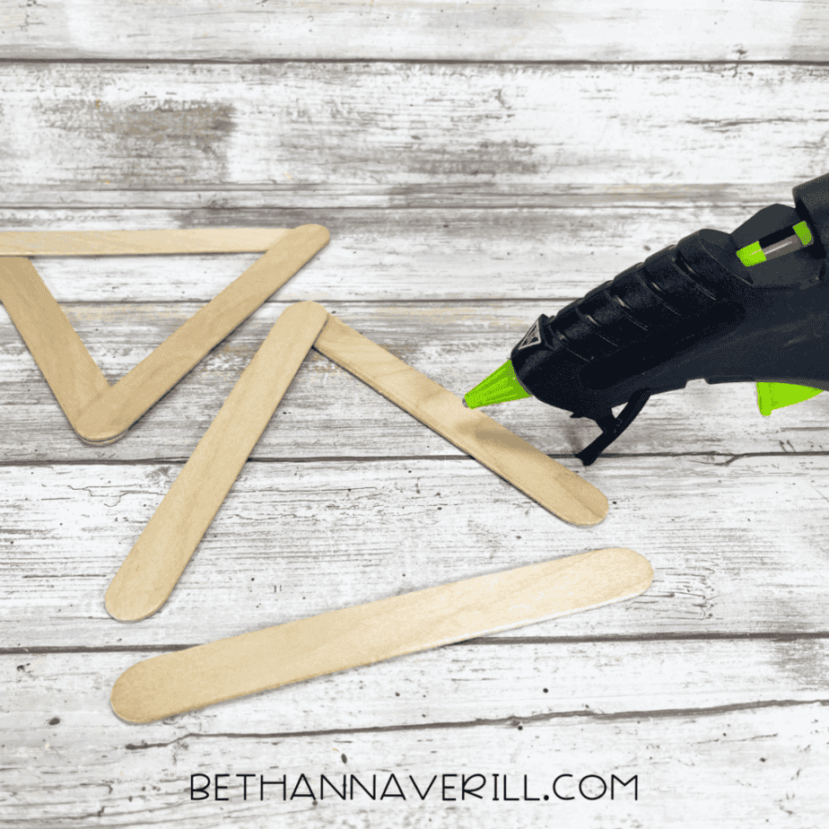 A hot glue gun assembling three craft sticks into a triangle shape on a wood-textured surface, with another finished triangle beside it.