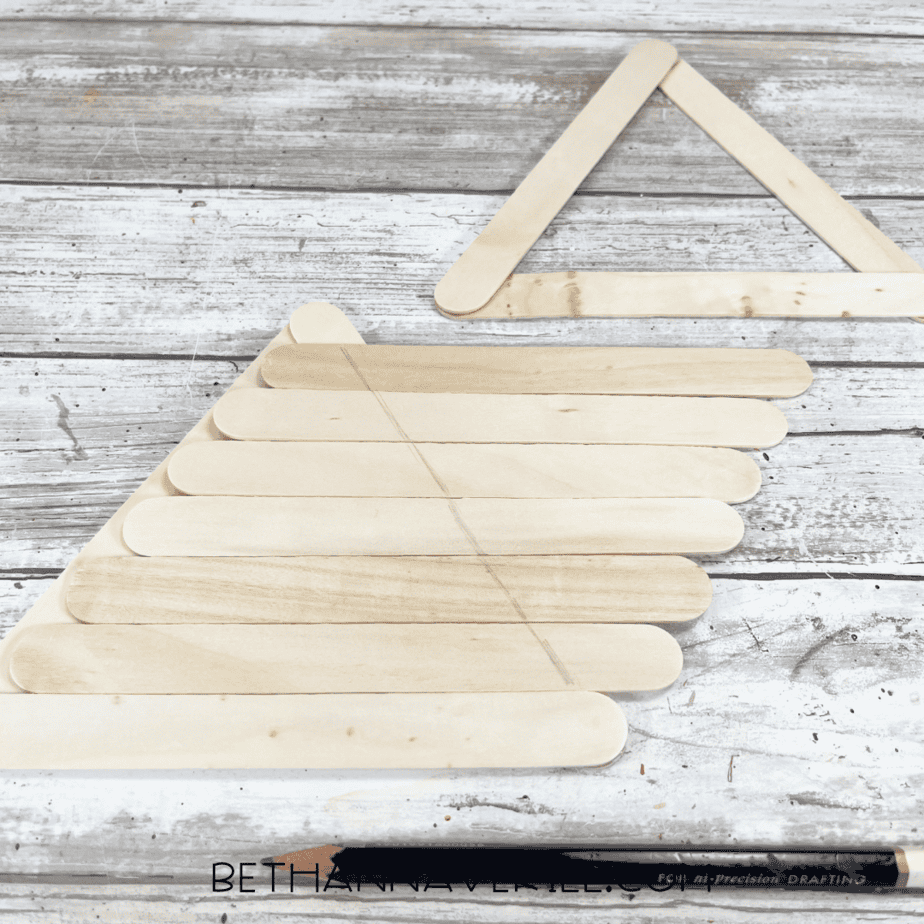 Several craft sticks laid out horizontally in a triangular shape, with pencil lines marking a cone outline to match a nearby popsicle stick triangle.