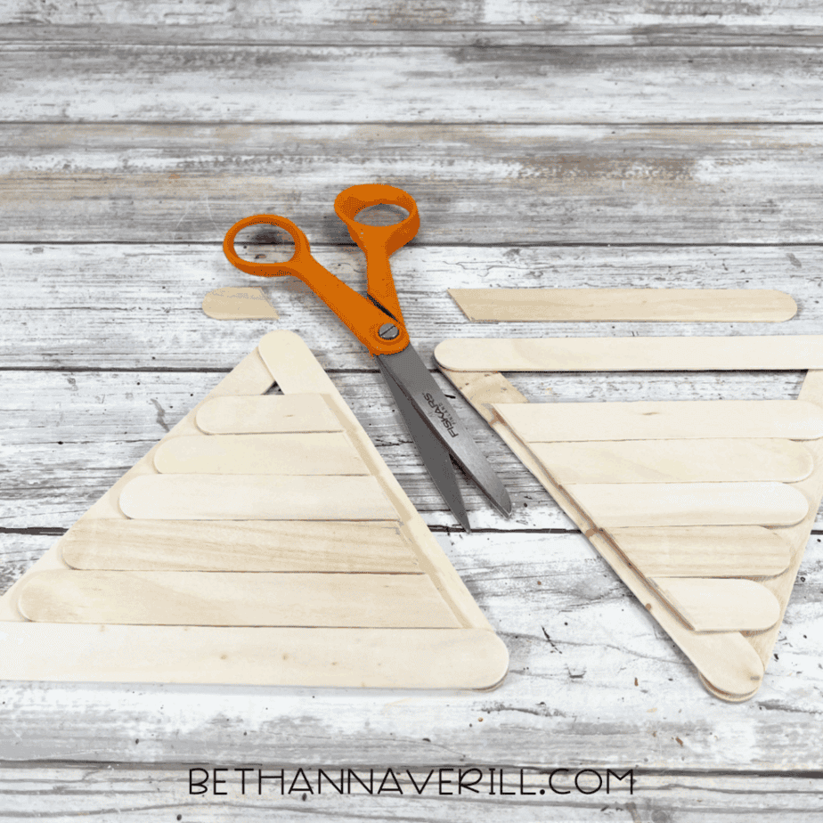 Two popsicle stick triangles filled with trimmed horizontal sticks, forming the base of an ice cream cone shape, with scissors laying nearby.