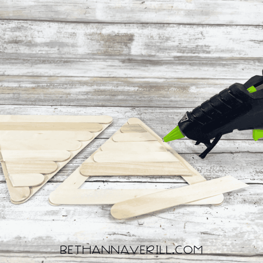 A glue gun being used to secure an extra craft stick along the border of a triangle-shaped popsicle stick cone, with one finished and one in progress.