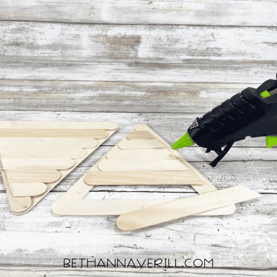 A glue gun being used to secure an extra craft stick along the border of a triangle-shaped popsicle stick cone, with one finished and one in progress.