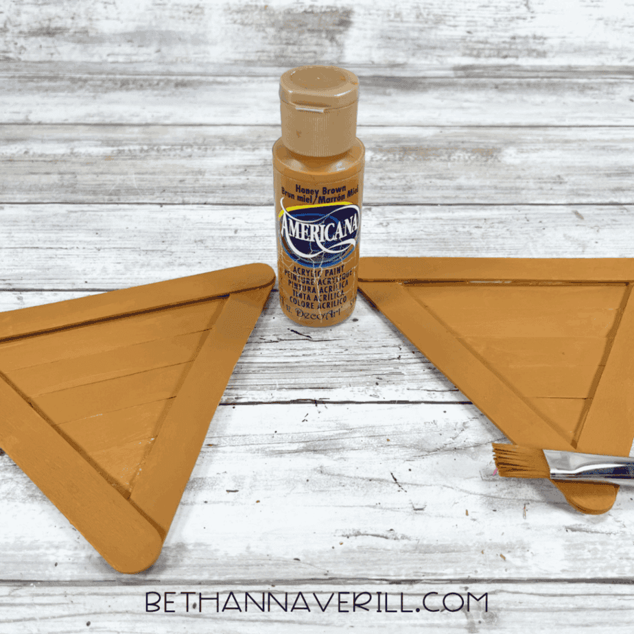 Two finished triangle-shaped cones painted a honey brown color, with a bottle of Americana acrylic paint and a paintbrush beside them.