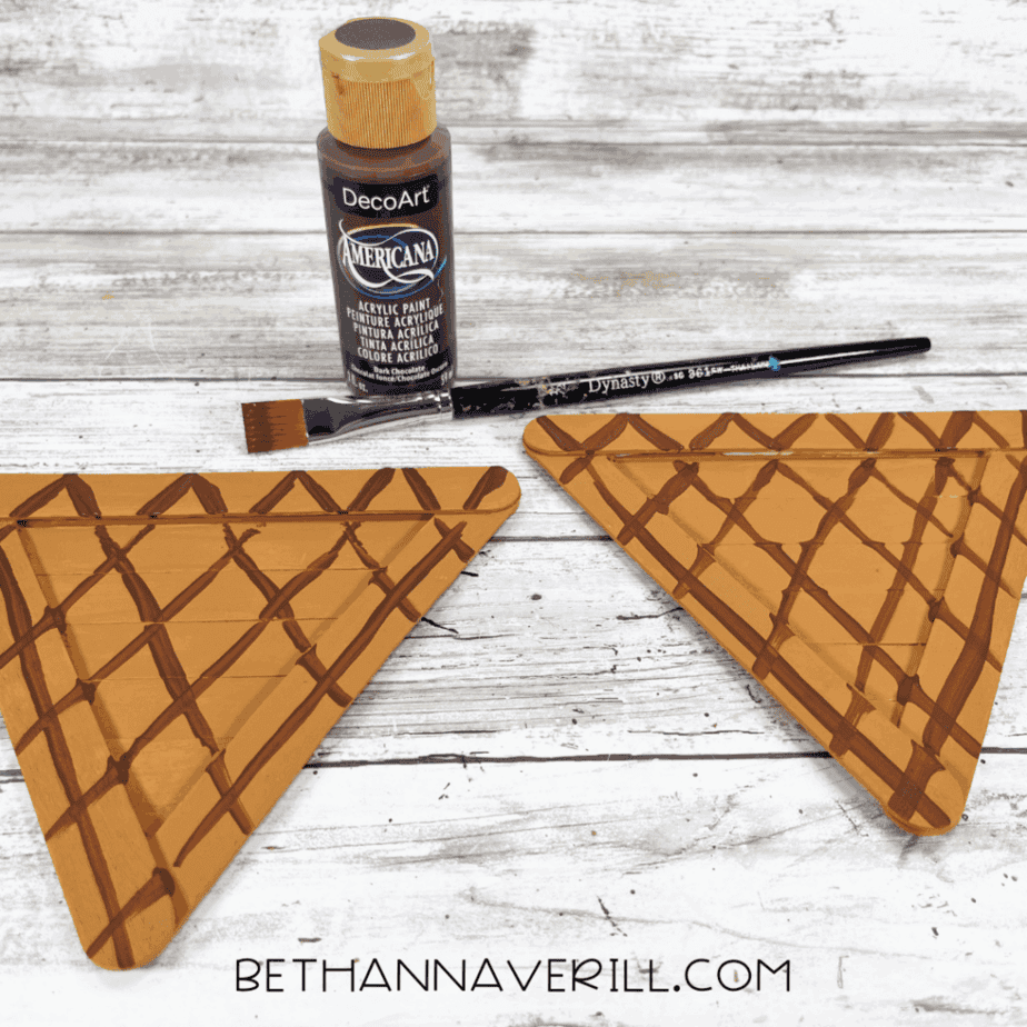 Two cone shapes painted with a crisscrossed chocolate brown pattern to mimic waffle cones, next to a bottle of dark brown acrylic paint and a brush.
