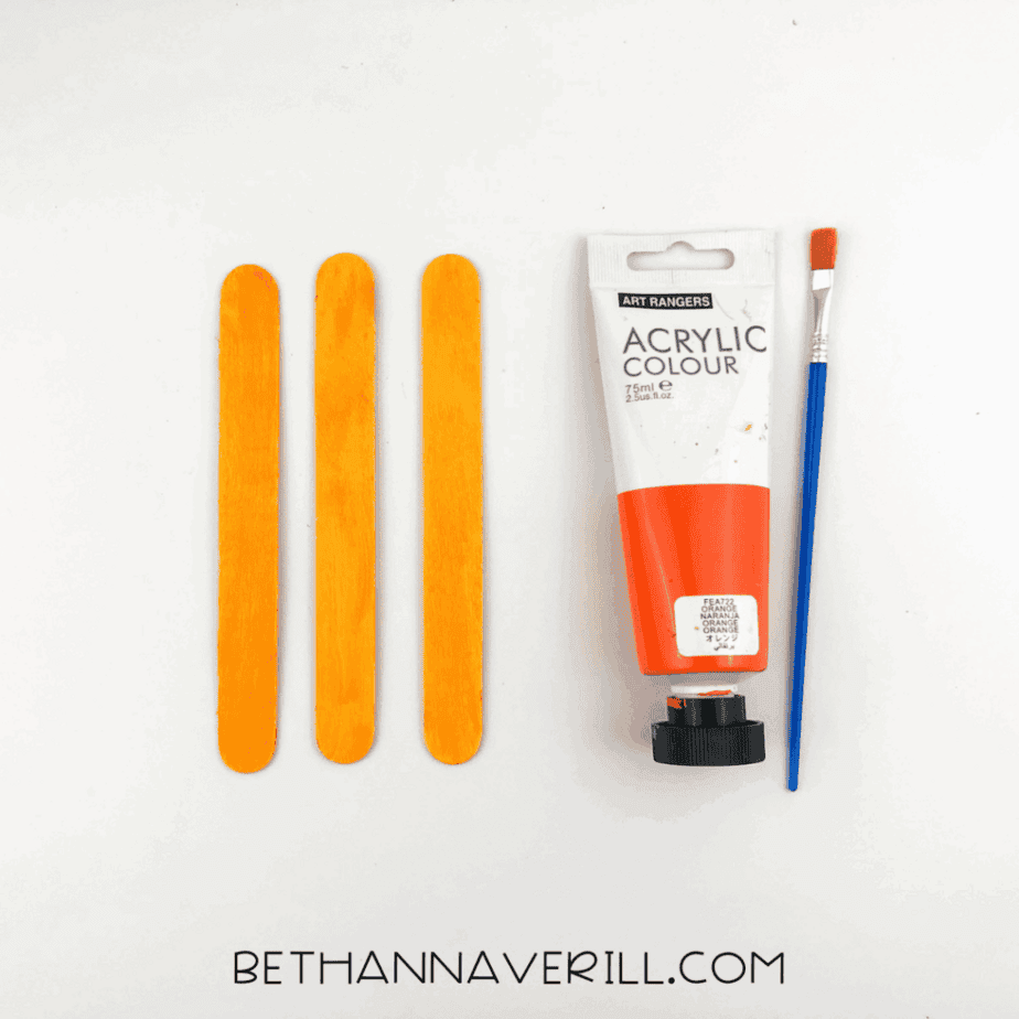 Three craft sticks painted orange next to orange paint and paintbrush.
