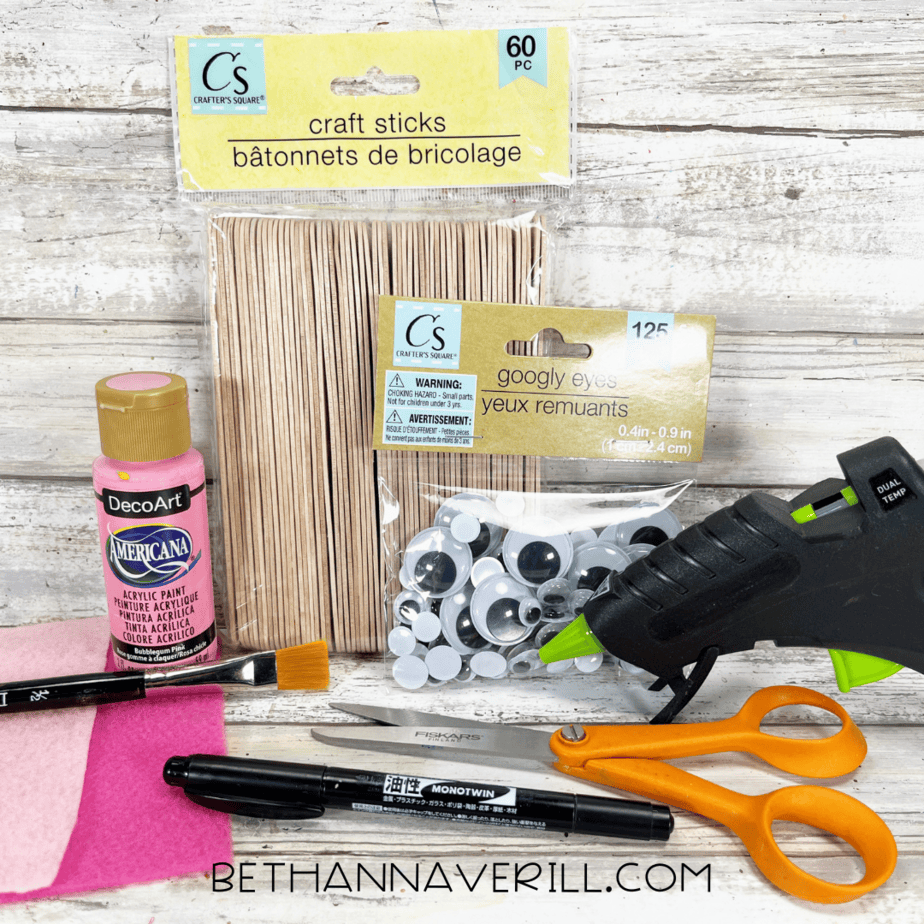 Pig popsicle stick craft supplies including craft sticks, pink paint, felt, googly eyes, marker, scissors, glue gun, and paintbrush.