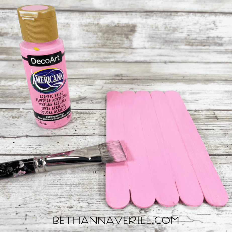 Craft stick base painted pink with paintbrush and bottle of pink paint next to it.