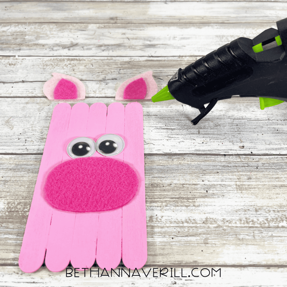 Pink painted craft stick base with googly eyes and a felt snout glued on, ears ready to attach.