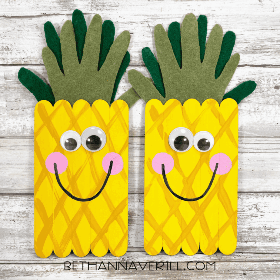 Completed pineapple popsicle stick craft — two finished pineapples with googly eyes, pink cheeks, big smiles, and layered felt leaves on a rustic wood background.