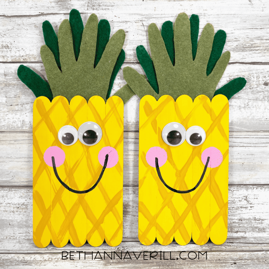 Completed pineapple popsicle stick craft — two finished pineapples with googly eyes, pink cheeks, big smiles, and layered felt leaves on a rustic wood background.
