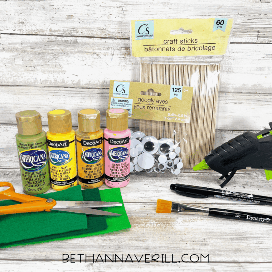 Materials laid out for a pineapple popsicle stick craft — yellow, light green, dark green, and pink acrylic paint, craft sticks, googly eyes, glue gun, scissors, green felt, paintbrush, and black marker on a rustic wood background.