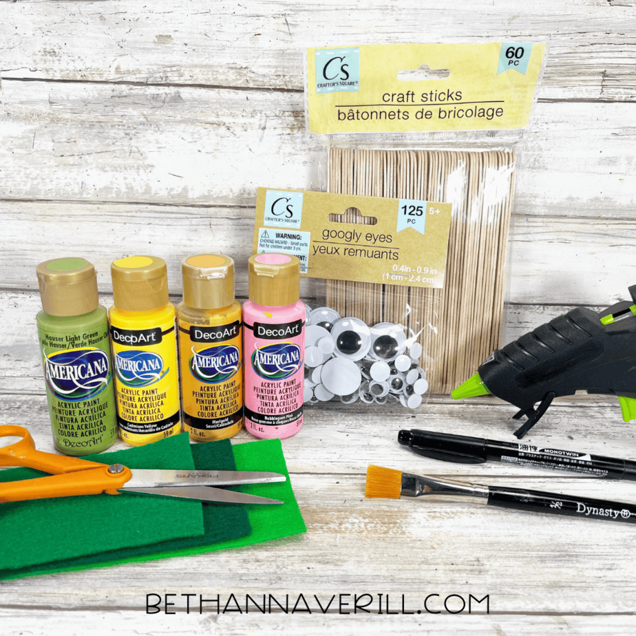 Materials laid out for a pineapple popsicle stick craft — yellow, light green, dark green, and pink acrylic paint, craft sticks, googly eyes, glue gun, scissors, green felt, paintbrush, and black marker on a rustic wood background.