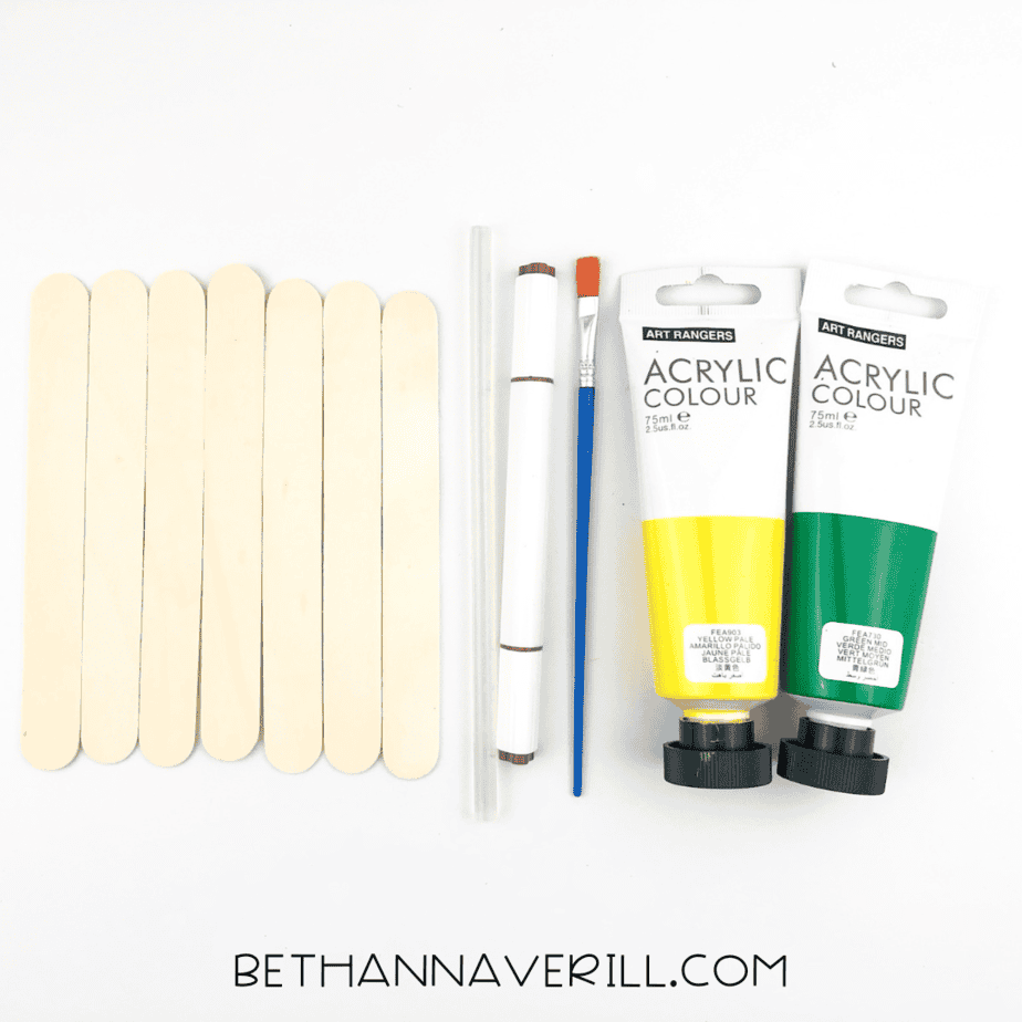 Craft materials laid out including popsicle sticks, paintbrush, yellow and green acrylic paint, and a brown marker.