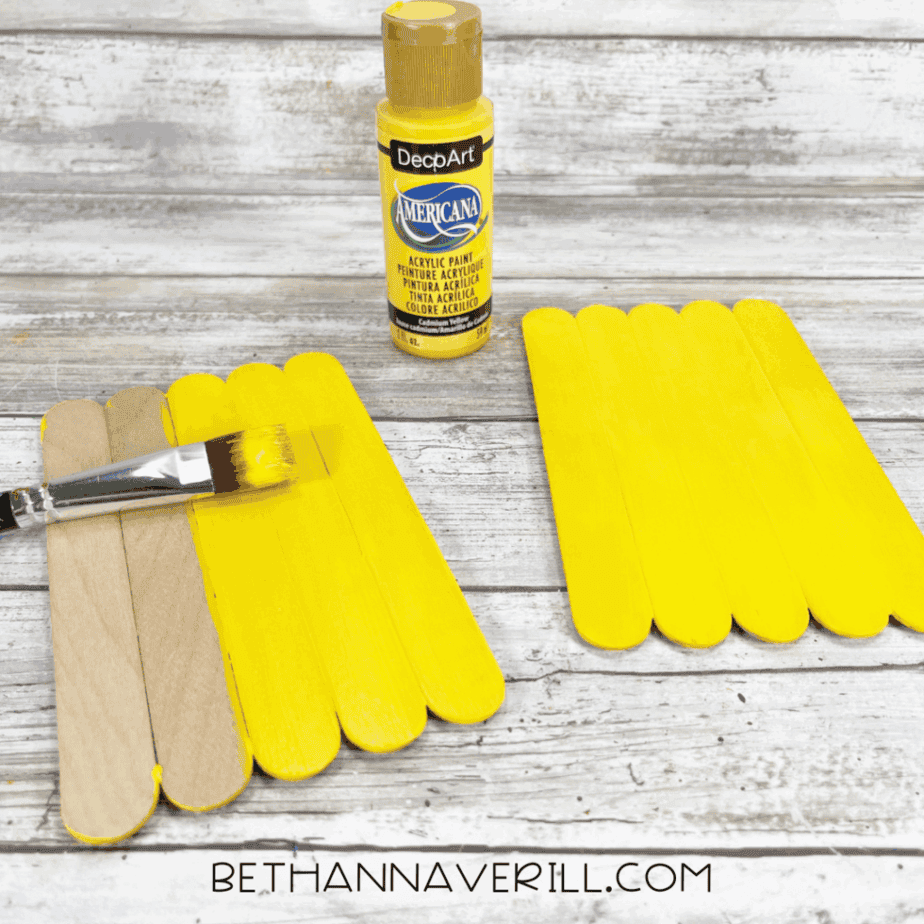 Painting the pineapple base yellow — flat paintbrush applying yellow acrylic paint to the craft stick panel.