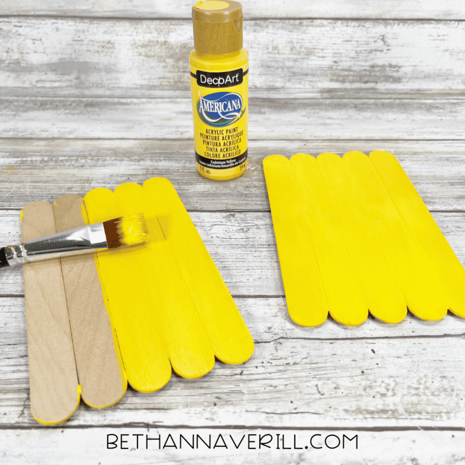 Painting the pineapple base yellow — flat paintbrush applying yellow acrylic paint to the craft stick panel.