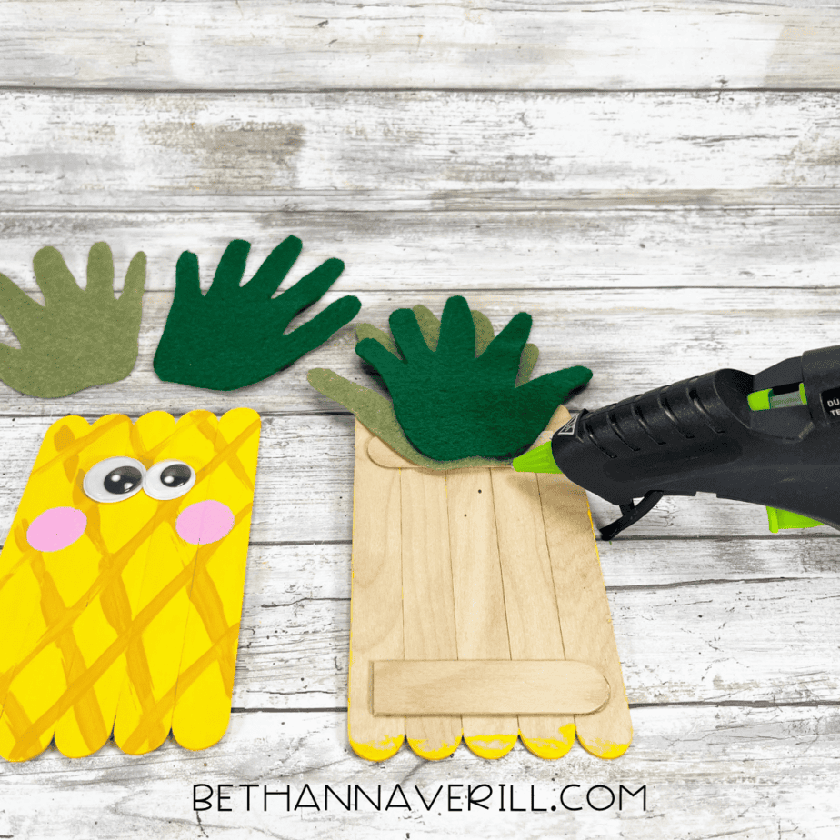 Gluing pineapple leaves to the back — hot glue gun attaching layered felt hand shapes to the top back of the pineapple base.