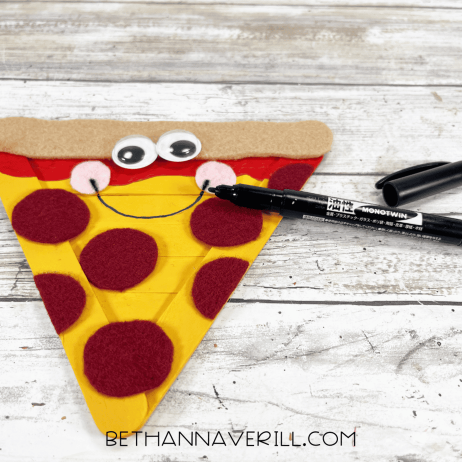 Drawing a smile — black marker adding a curved smile below the eyes and cheeks on the pizza face.