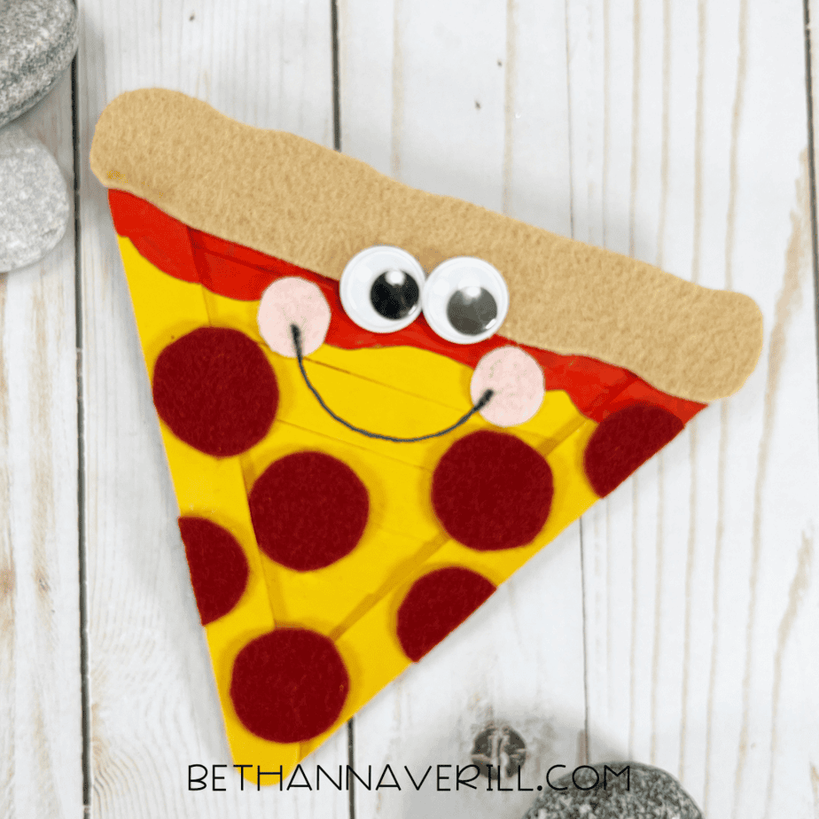 Finished pizza slice craft — completed smiling pizza slice with googly eyes, pink felt cheeks, and red felt pepperoni, displayed on a light wood background with decorative rocks.