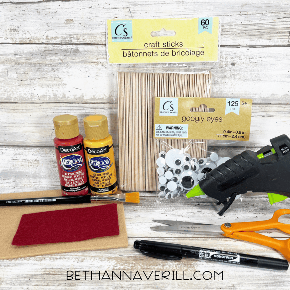 Materials laid out for a pizza popsicle stick craft — red and yellow acrylic paint, craft sticks, googly eyes, glue gun, scissors, felt sheets, paintbrush, and black marker on a rustic wood background.