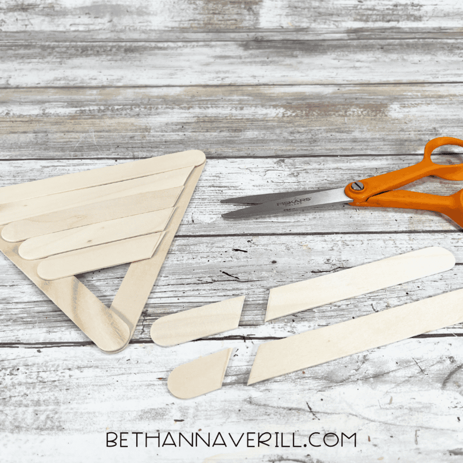 Trimming excess craft sticks — scissors cutting the overhanging craft stick edges to fit the triangle shape.