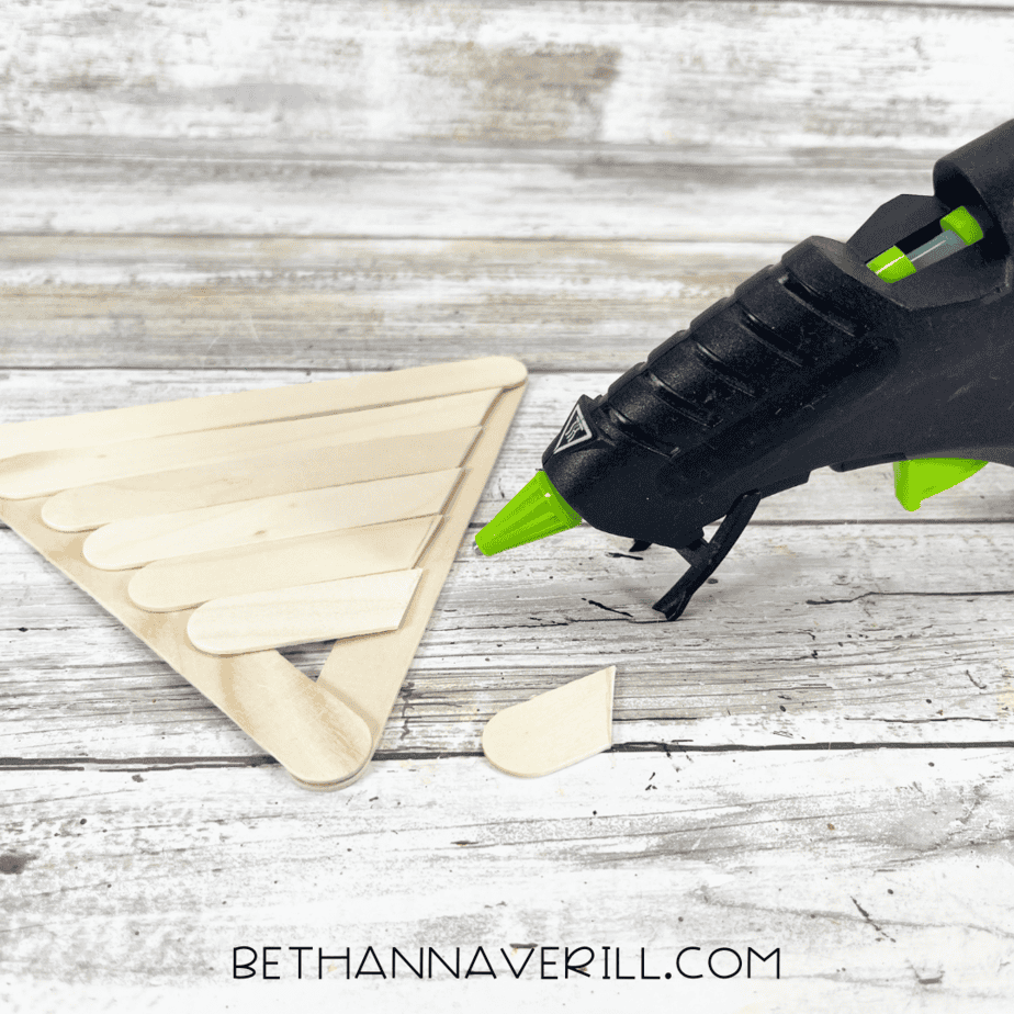 Gluing trimmed craft sticks into place — hot glue gun attaching the trimmed pieces to complete the pizza slice base.