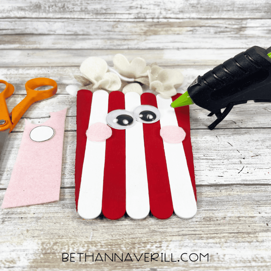 A partially assembled popcorn container made from red and white craft sticks with felt popcorn on top, googly eyes, and pink felt cheeks next to a hot glue gun and scissors.