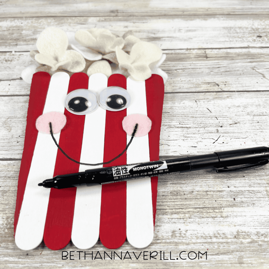 A black marker is used to draw a smile on the face of the popcorn craft, which now has googly eyes and pink cheeks.