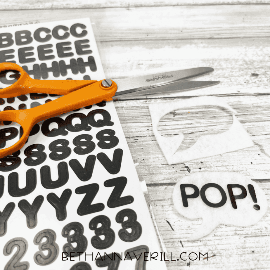 A pair of scissors sits next to a sheet of black alphabet stickers and a white felt speech bubble that reads “POP!”