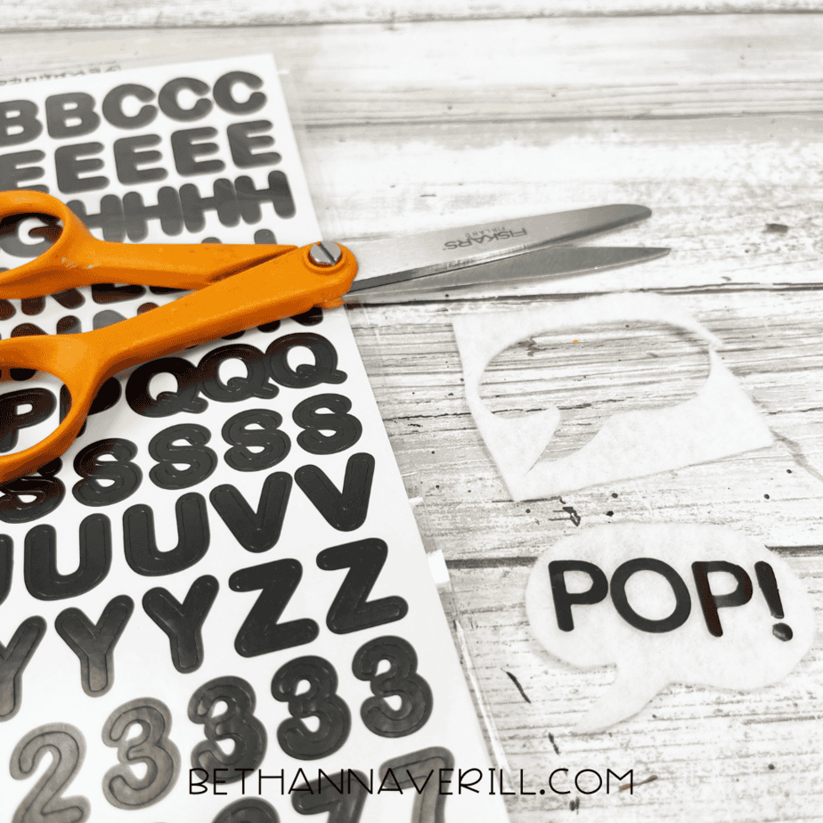 A pair of scissors sits next to a sheet of black alphabet stickers and a white felt speech bubble that reads “POP!”