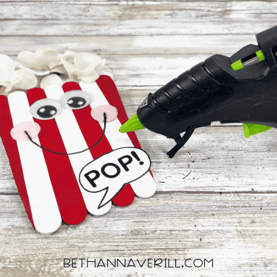A glue gun is used to attach the speech bubble with the word “POP!” to the bottom of the popcorn craft.