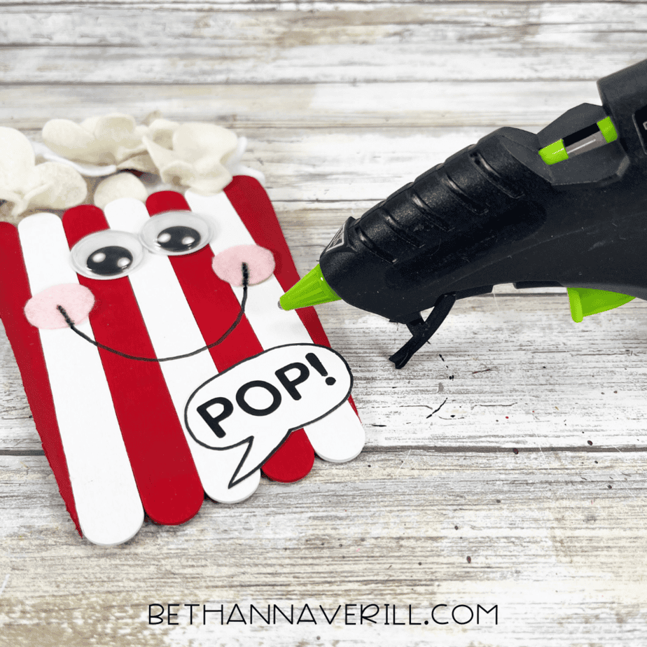 A glue gun is used to attach the speech bubble with the word “POP!” to the bottom of the popcorn craft.