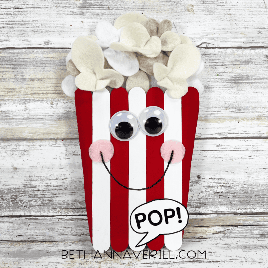 Finished popcorn craft with a big smile, rosy cheeks, googly eyes, fluffy felt popcorn, and a “POP!” speech bubble.