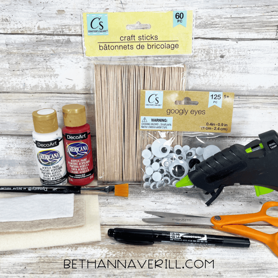 Craft materials laid out on a wood background, including acrylic paint, craft sticks, googly eyes, felt sheets, scissors, glue gun, brush, and black marker.
