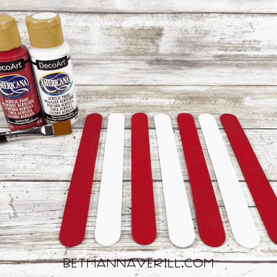 Eight craft sticks painted alternately red and white, laid out in a popcorn box stripe pattern next to red and white paint bottles.