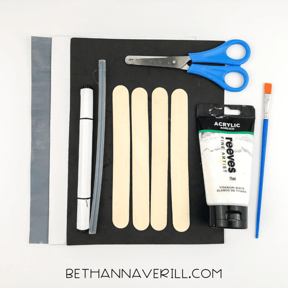 Supplies for raccoon popsicle stick craft including four sticks, white paint, brush, scissors, black marker, and gray, white, and black paper.