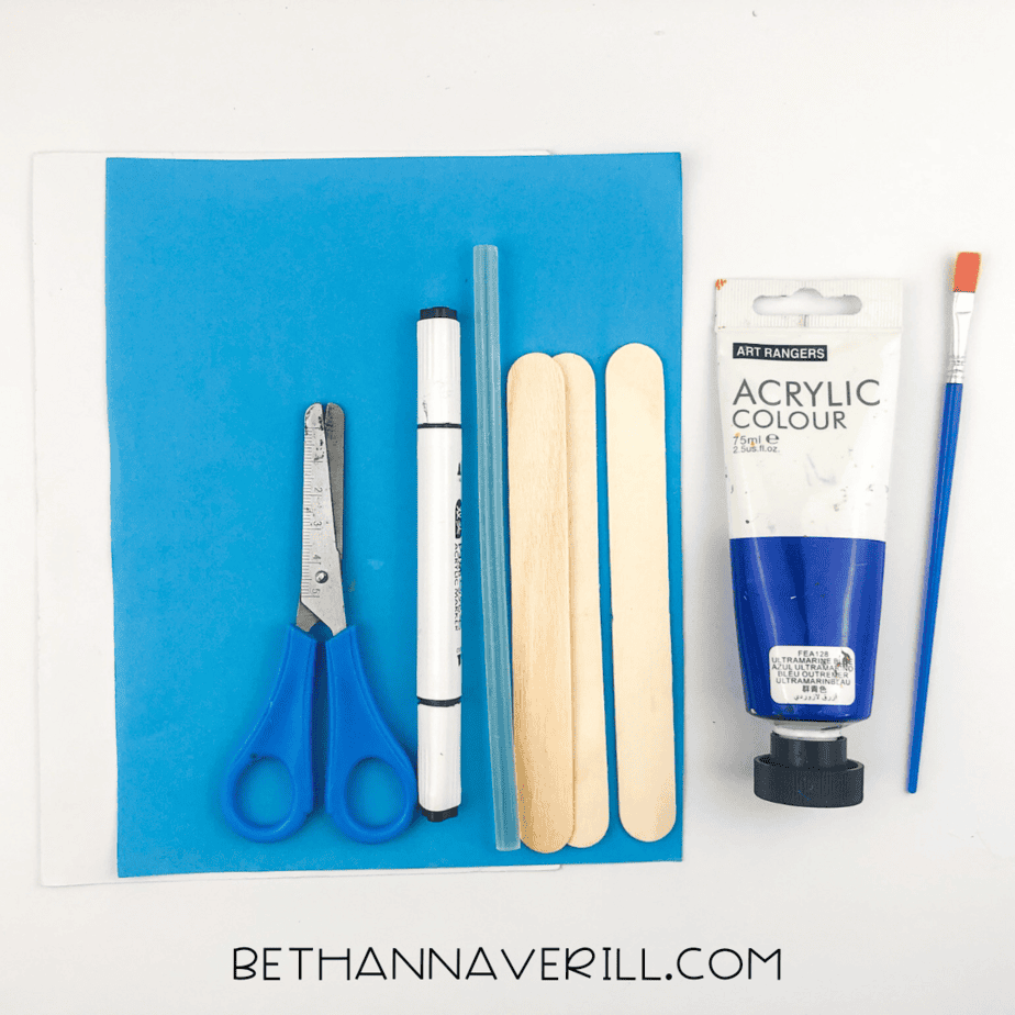 Shark craft supplies laid out: blue acrylic paint, paintbrush, three jumbo craft sticks, blue construction paper, white paper, scissors, glue, and a black marker.