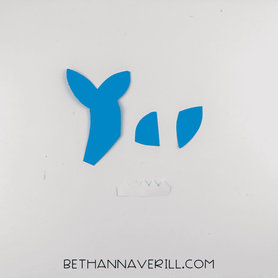 Blue paper shapes cut for the shark’s tail, dorsal fin, and side fin, along with white paper teeth and an eye.