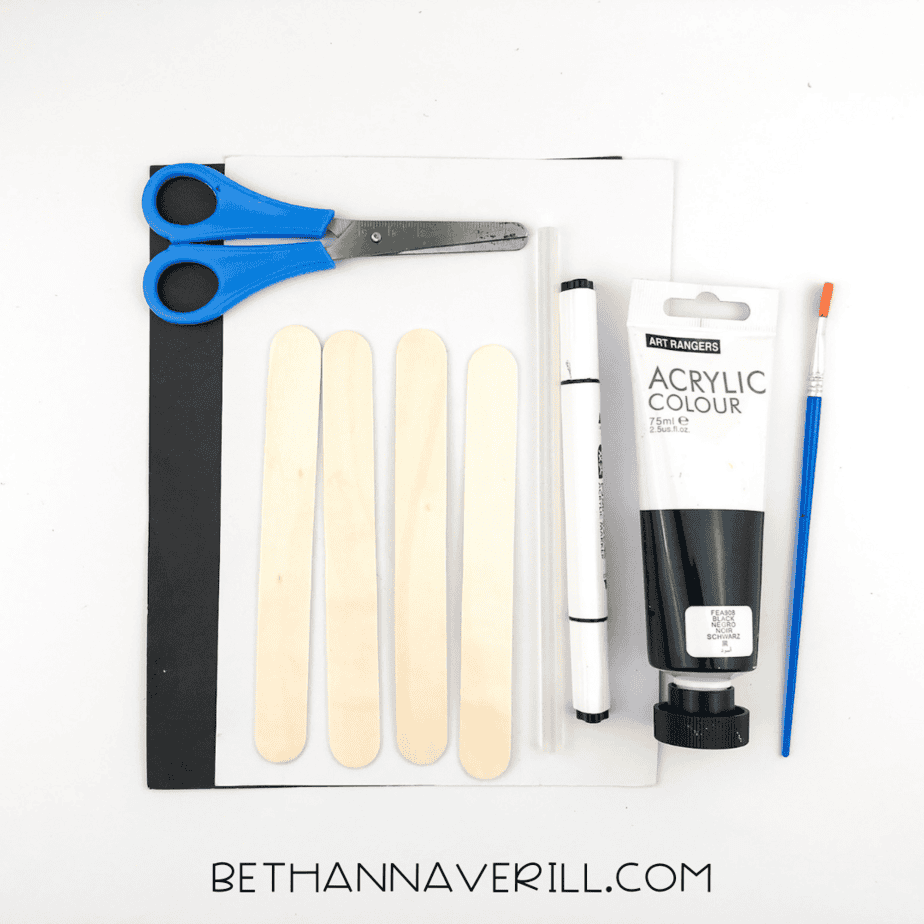 Flat‑lay of supplies: four plain craft sticks, black and white paper, scissors, glue sticks, black acrylic paint, paintbrush, and a marker.