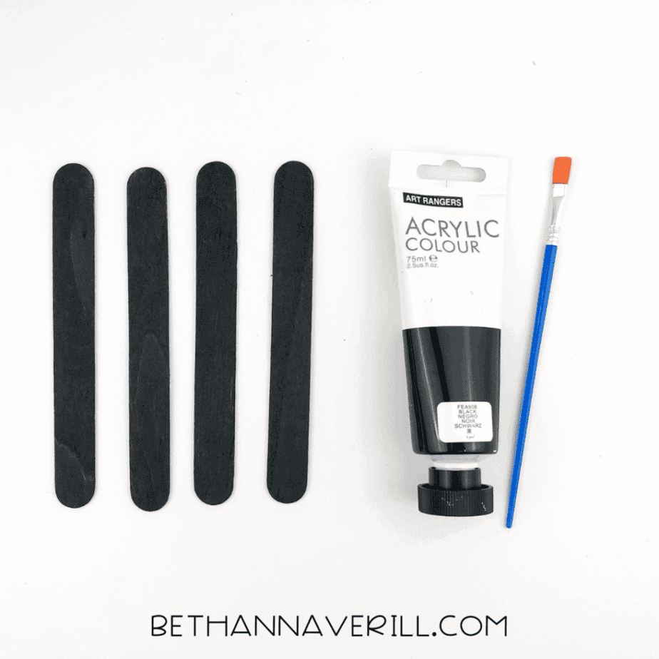 Four craft sticks painted black drying beside a tube of black acrylic paint and a brush.