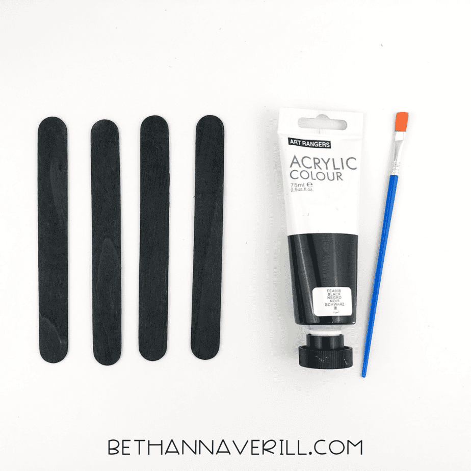 Four craft sticks painted black drying beside a tube of black acrylic paint and a brush.