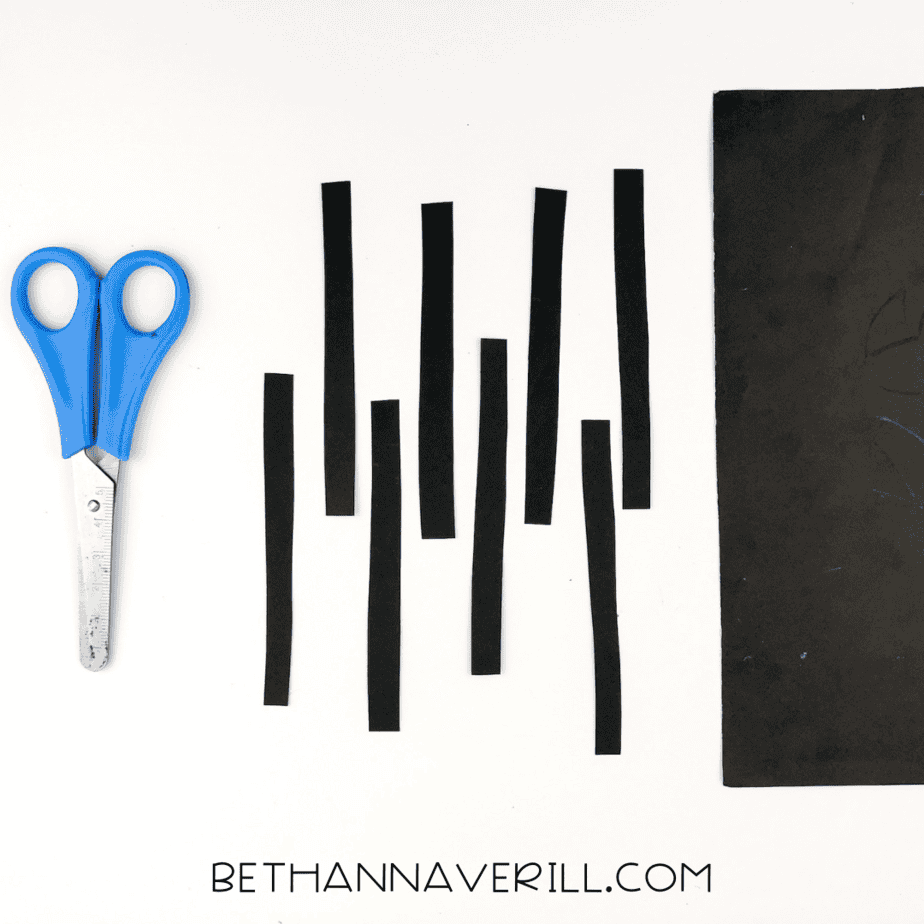 Thin strips of black construction paper cut for spider legs next to scissors and paper.