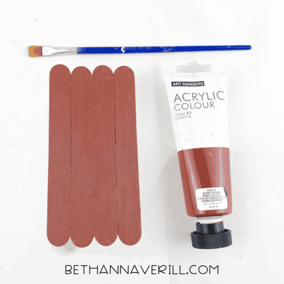 Painted brown craft sticks next to a paintbrush and acrylic paint.
