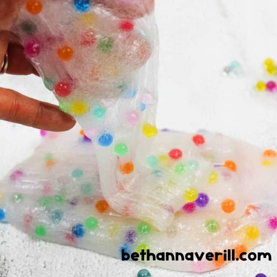 Fingers pull clear slime embedded with colorful water beads across a textured white surface, showing the slime’s elasticity and playful appearance.